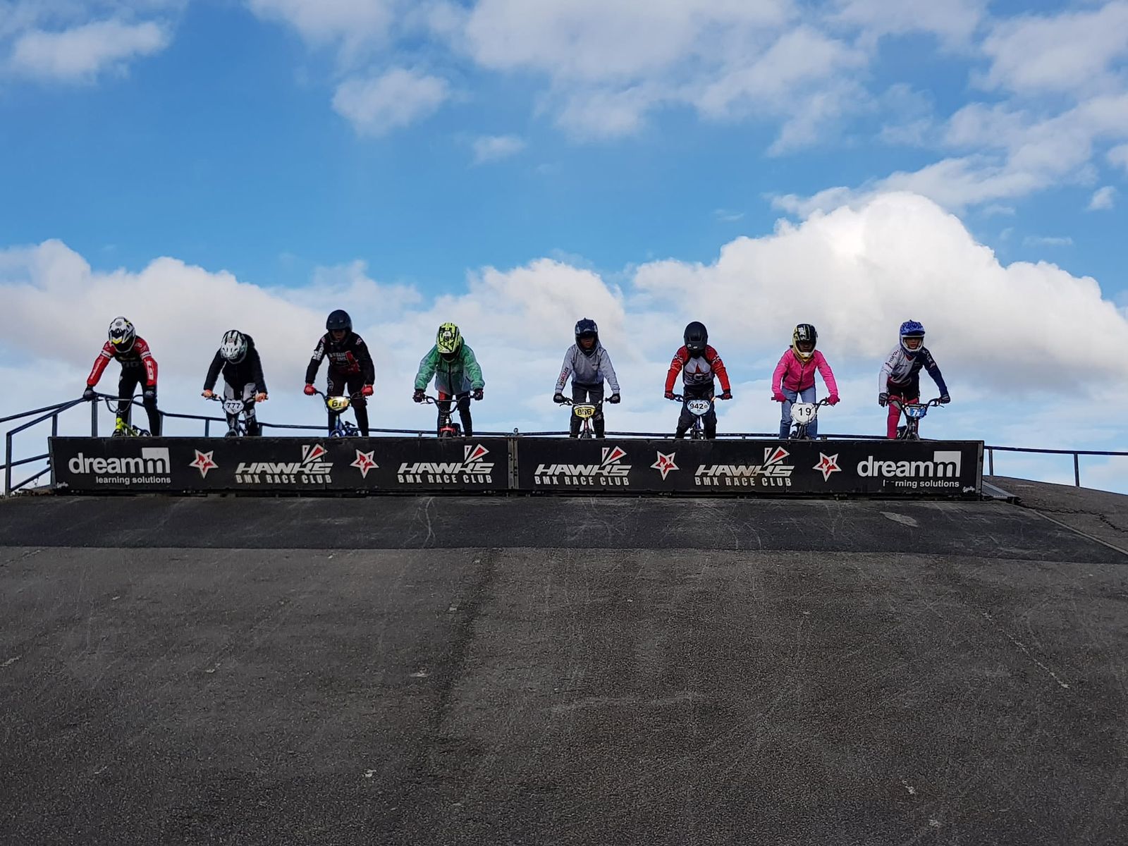 BMX riders lined up at the top of the start ramp against a blue sky